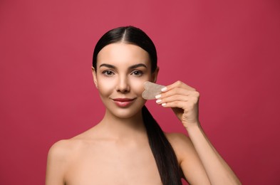 Photo of Beautiful young woman doing facial massage with gua sha tool on pink background