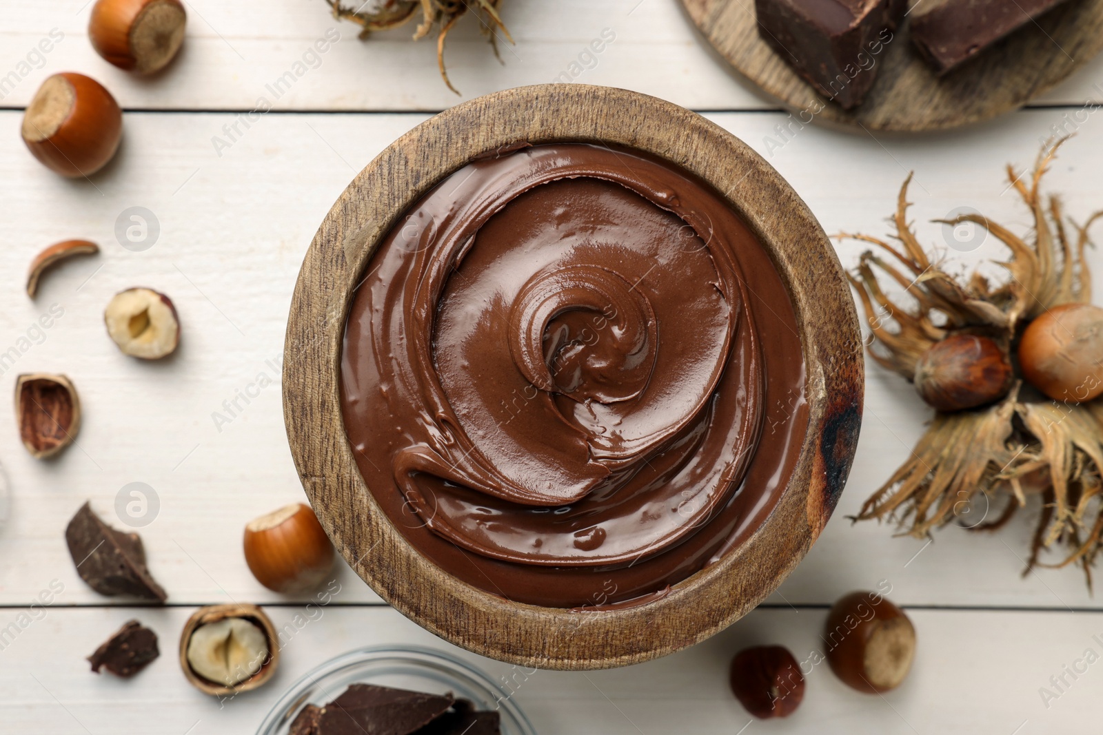 Photo of Bowl of tasty chocolate paste with hazelnuts on white wooden table, flat lay