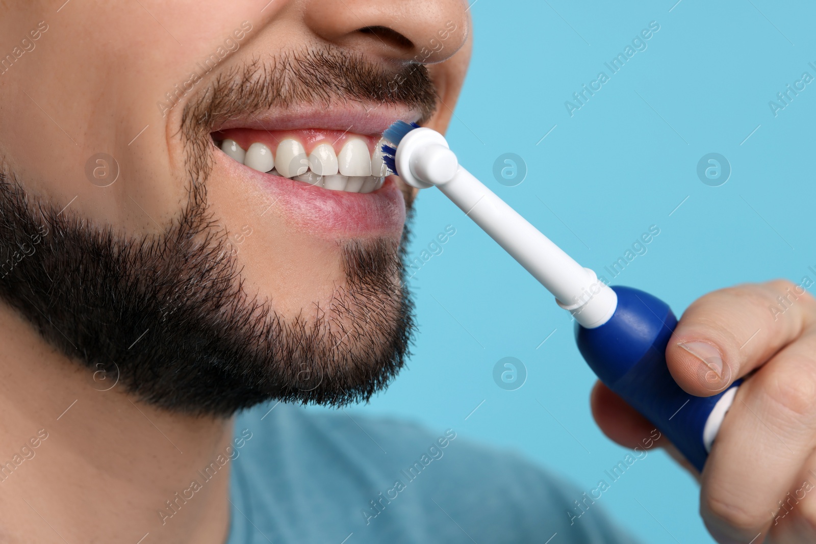 Photo of Man brushing his teeth with electric toothbrush on light blue background, closeup