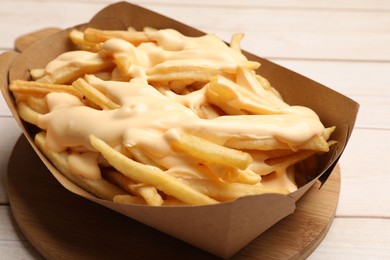 Tasty potato fries and cheese sauce in paper container on light wooden table, closeup