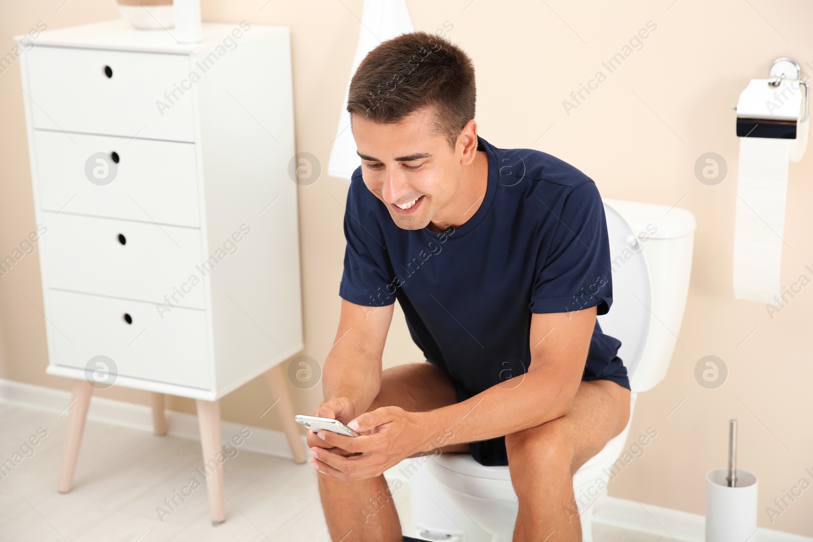 Photo of Young man using mobile phone while sitting on toilet bowl at home