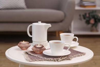 Photo of Cups of tea, teapot and burning candles on white coffee table indoors