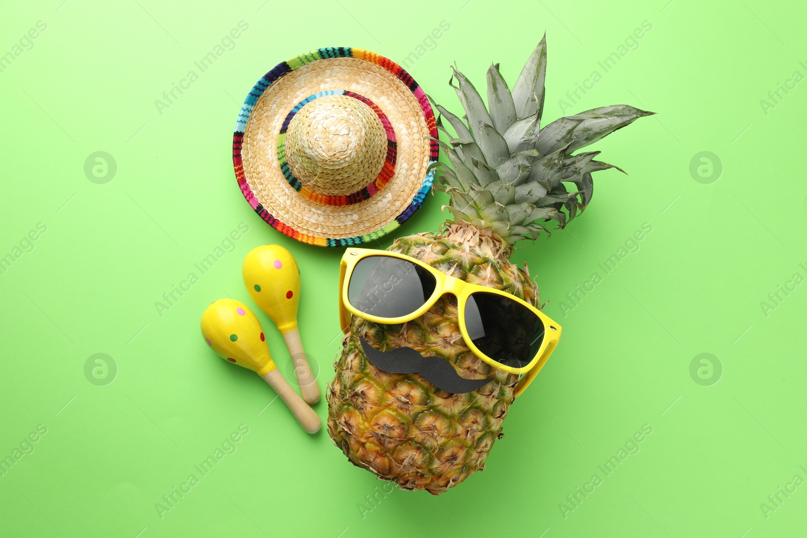 Photo of Mexican sombrero hat, pineapple with sunglasses, fake mustache and maracas on green background, flat lay