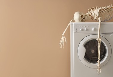 Waiting concept. Human skeleton lying on washing machine near beige wall indoors, space for text