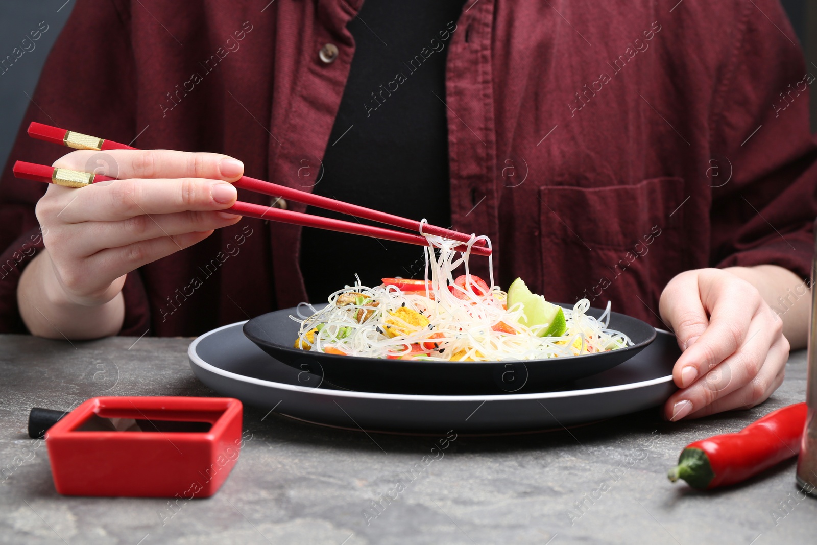 Photo of Stir-fry. Woman with chopsticks eating tasty rice noodles with meat and vegetables at grey textured table, closeup