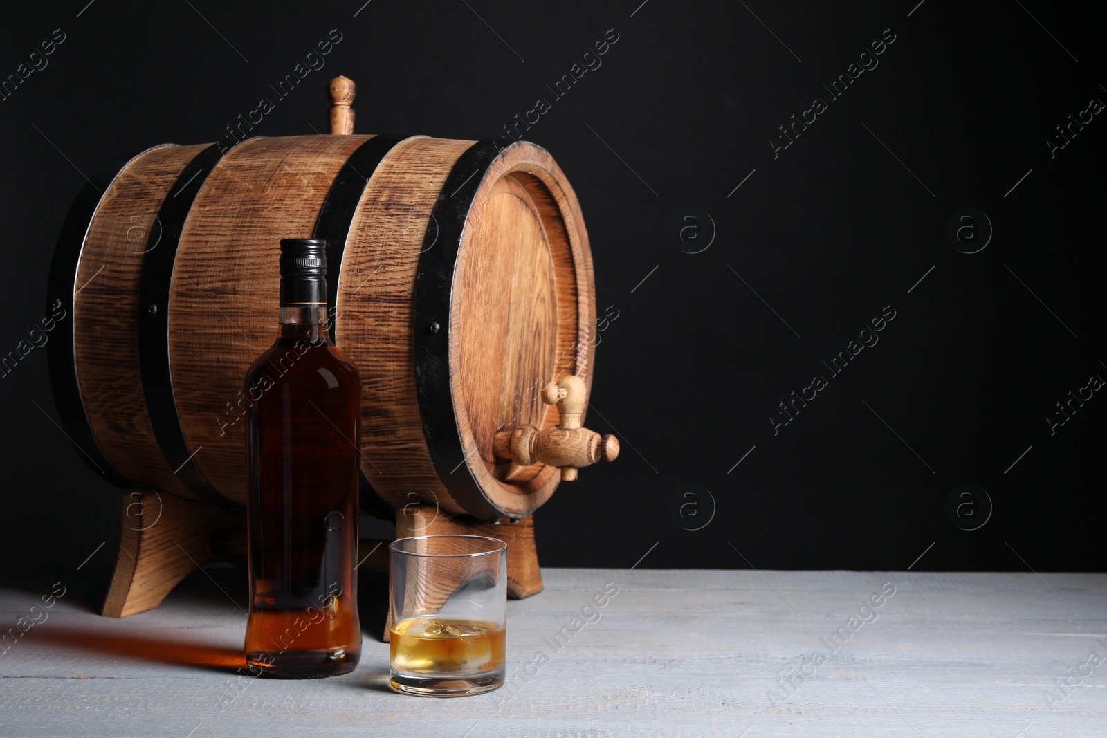 Photo of Barrel with tap, bottle and glass of tasty whiskey on light gray wooden table, space for text