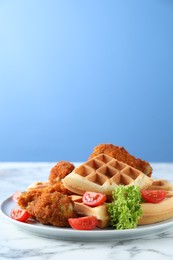 Photo of Tasty Belgian waffles served with fried chicken, tomatoes and lettuce on white marble table against light blue background, closeup. Space for text