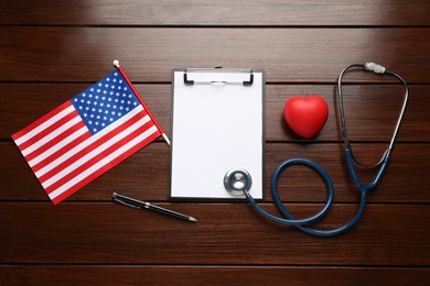 Photo of Stethoscope, red heart, clipboard and American flag on wooden table, flat lay