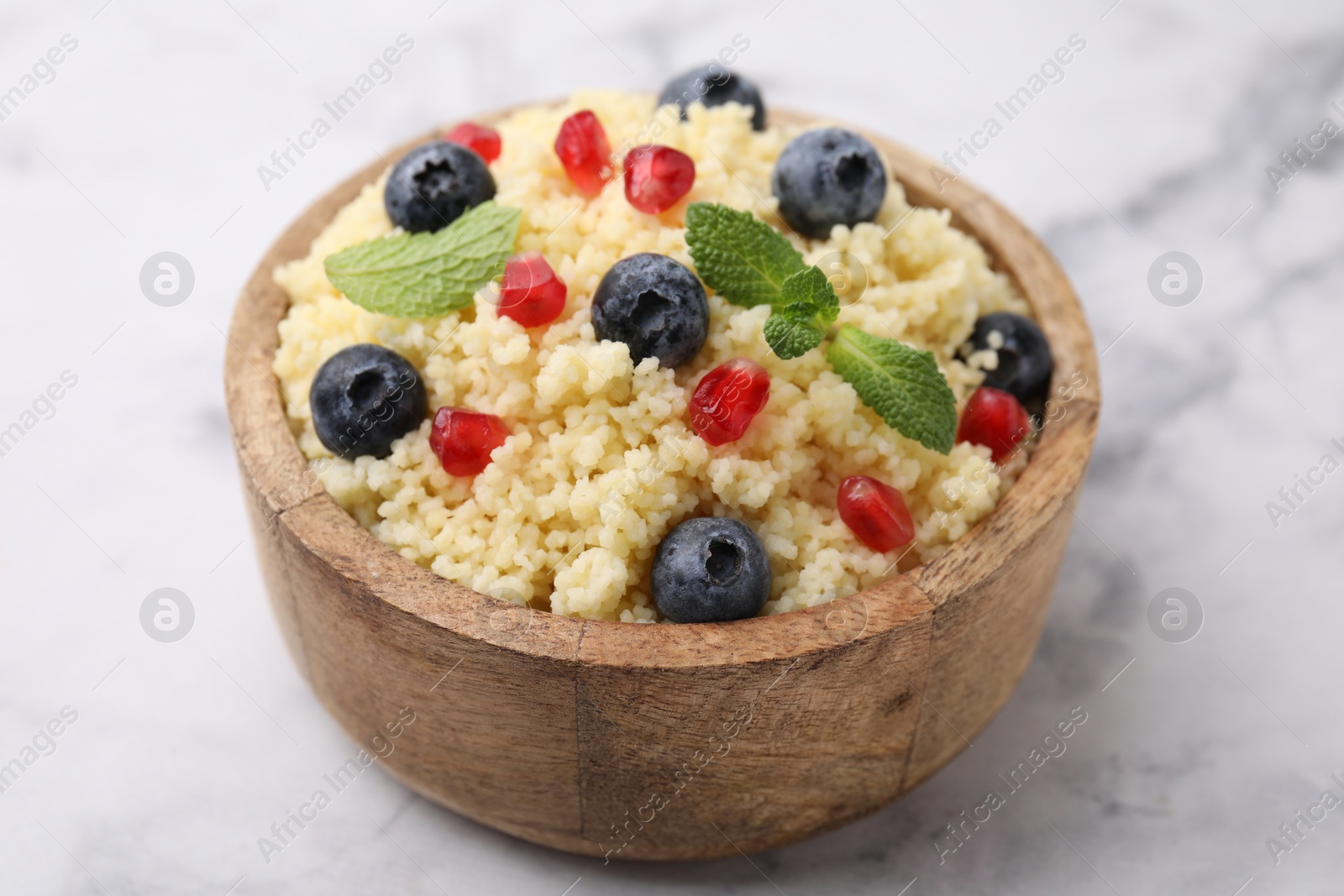 Photo of Bowl of tasty couscous with blueberries, pomegranate and mint on white marble table, closeup