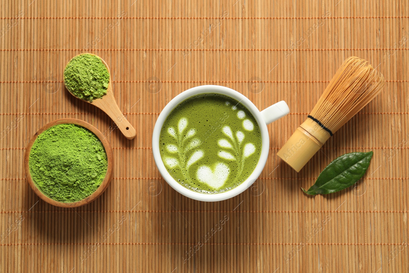Photo of Delicious matcha latte, powder, leaf and whisk on bamboo mat, flat lay