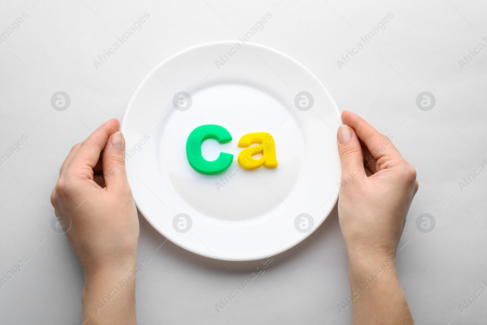 Photo of Woman holding plate with calcium symbol made of colorful letters on light grey background, top view