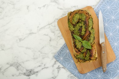 Freshly baked pesto bread with basil and knife on white marble table, top view. Space for text