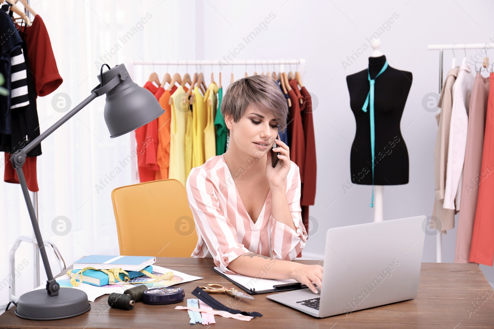 Photo of Beautiful young stylist talking on phone at workplace near rack with clothes