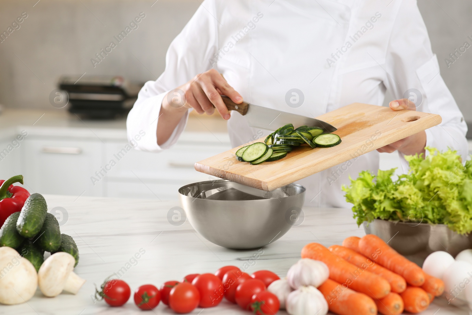 Photo of Professional chef putting cut cucumber into metal bowl at white marble table in kitchen, closeup