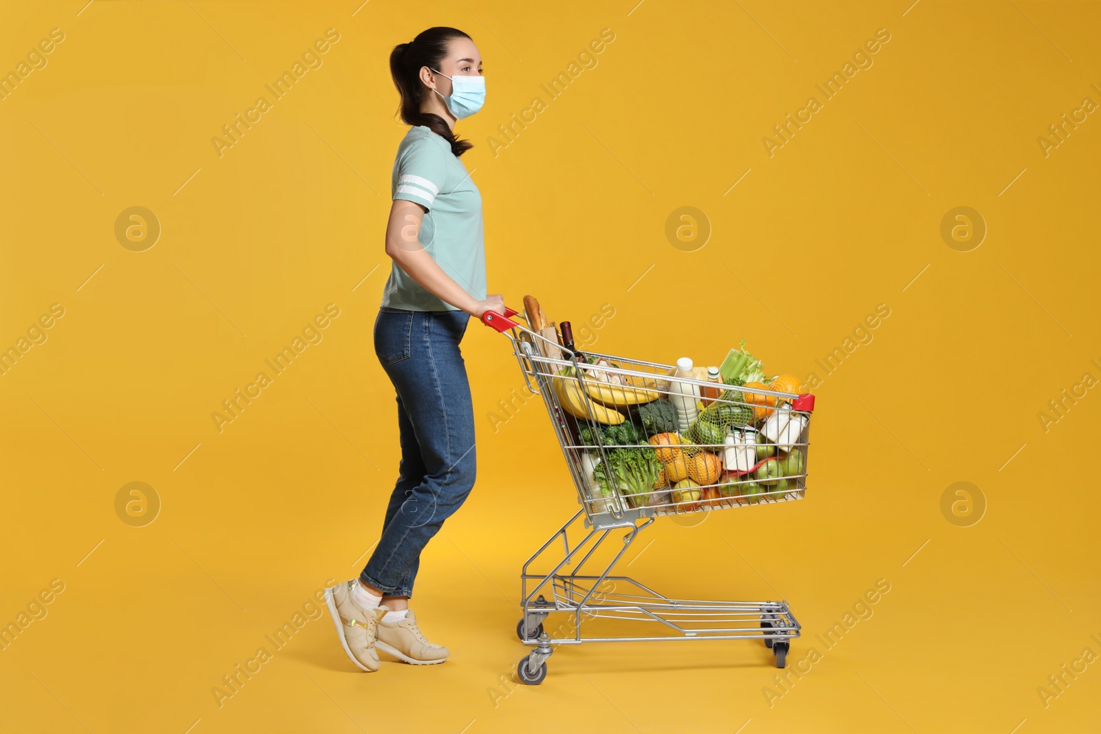 Photo of Woman with protective mask and shopping cart full of groceries on yellow background