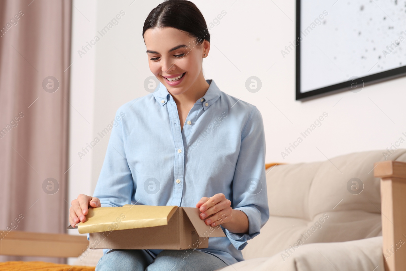 Photo of Happy young woman opening parcel on sofa at home. Internet shopping