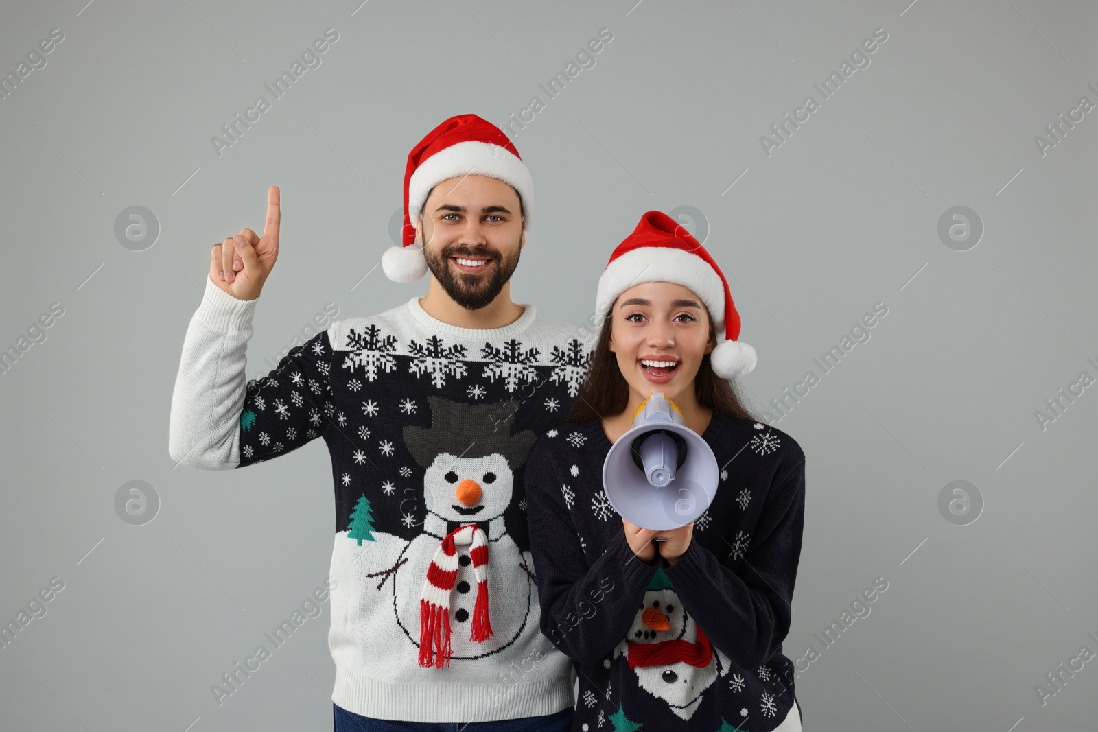 Photo of Young couple in Christmas sweaters and Santa hats shouting in megaphone on grey background
