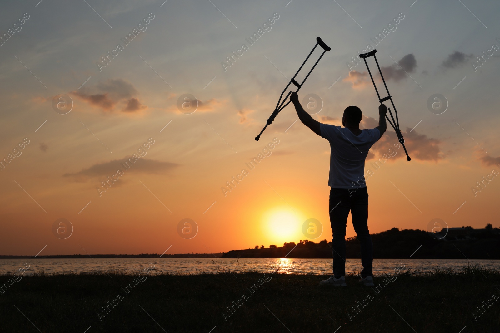 Photo of Man raising hands with underarm crutches up to sky near river at sunset, back view. Healing miracle