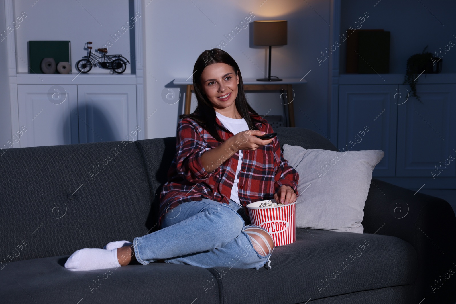 Photo of Happy woman holding popcorn bucket and changing TV channels with remote control at home in evening