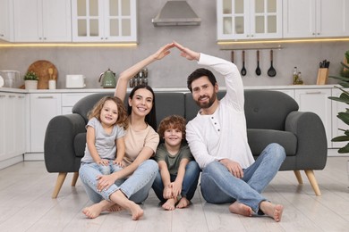 Photo of Family housing concept. Happy woman and her husband forming roof with their hands while sitting with kids on floor at home