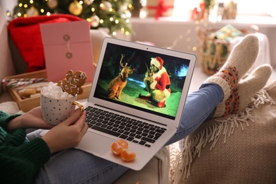 Photo of MYKOLAIV, UKRAINE - DECEMBER 25, 2020: Woman with sweet drink watching The Grinch movie on laptop at home, closeup. Cozy winter holidays atmosphere
