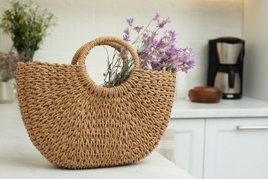 Stylish beach bag with beautiful bouquet of wildflowers on white table in kitchen. Space for text