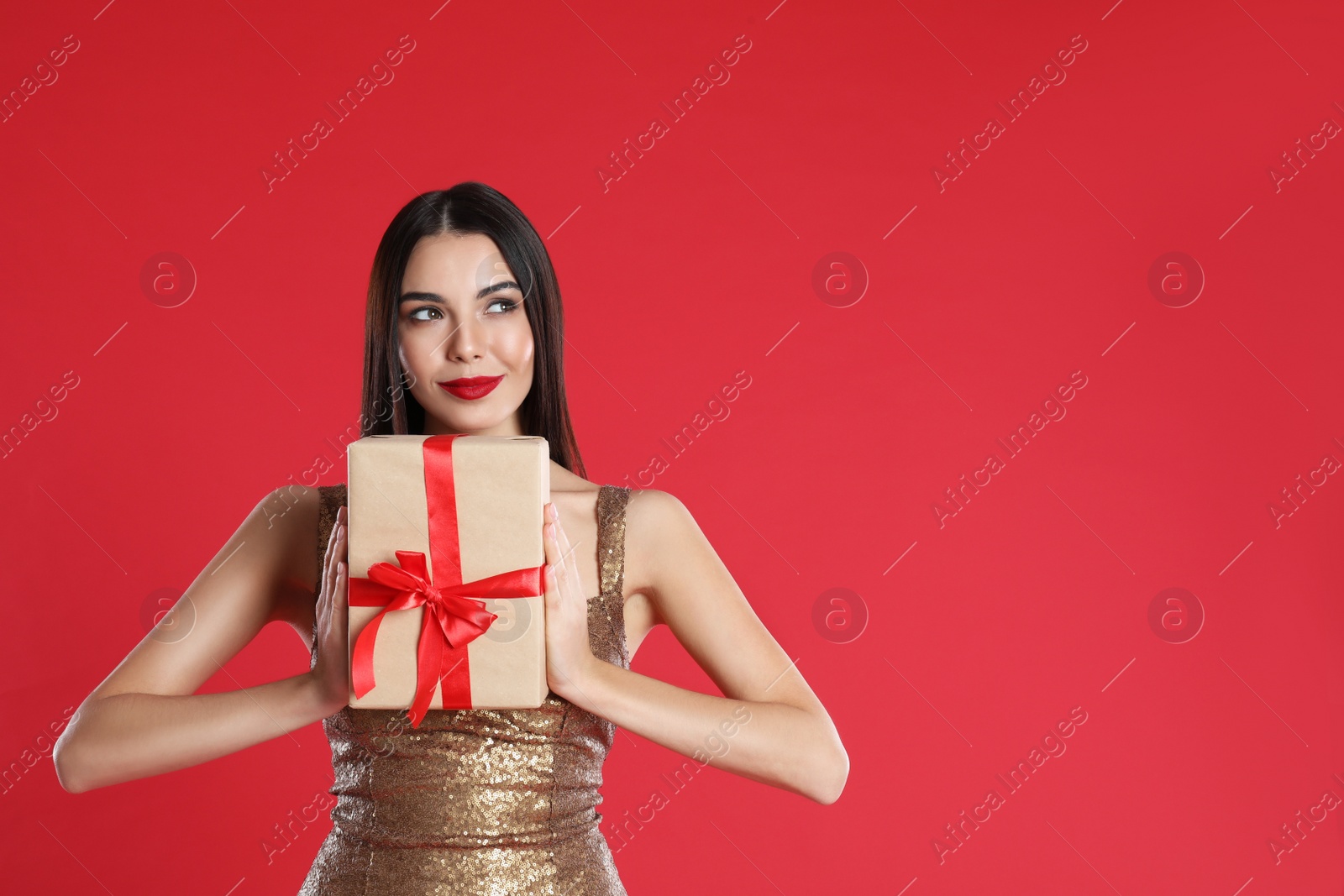 Photo of Woman in golden dress holding Christmas gift on red background, space for text