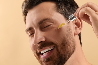 Photo of Smiling man applying cosmetic serum onto his face on beige background, closeup