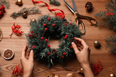 Photo of Florist making beautiful Christmas wreath with berries and pine cones at wooden table, above view