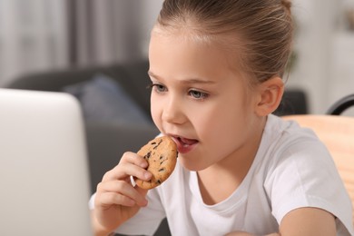 Photo of Little girl using laptop and eating biscuit at home. Internet addiction