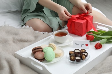 Photo of Tasty breakfast served in bed. Woman with gift box, desserts, tea, flowers and I Love You card at home, closeup