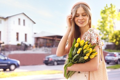 Beautiful teenage girl with bouquet of yellow tulips on city street, space for text