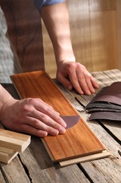 Photo of Man polishing wooden plank with sandpaper at table indoors, closeup