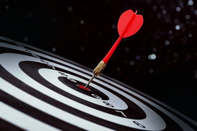 Image of Dart board with red arrow hitting target against black background, bokeh effect