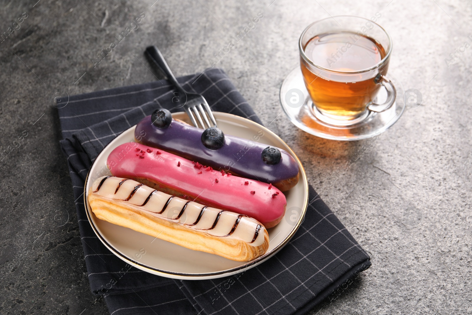 Photo of Different tasty glazed eclairs served with tea on grey textured table