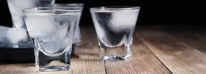 Shot glasses of vodka with ice cubes on wooden table against black background, closeup view with space for text. Banner design