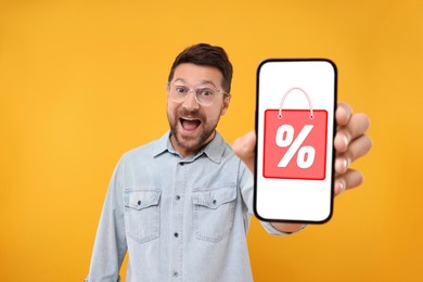 Image of Discount, offer, sale. Emotional man showing mobile phone with illustration of bag and percent sign on screen, orange background