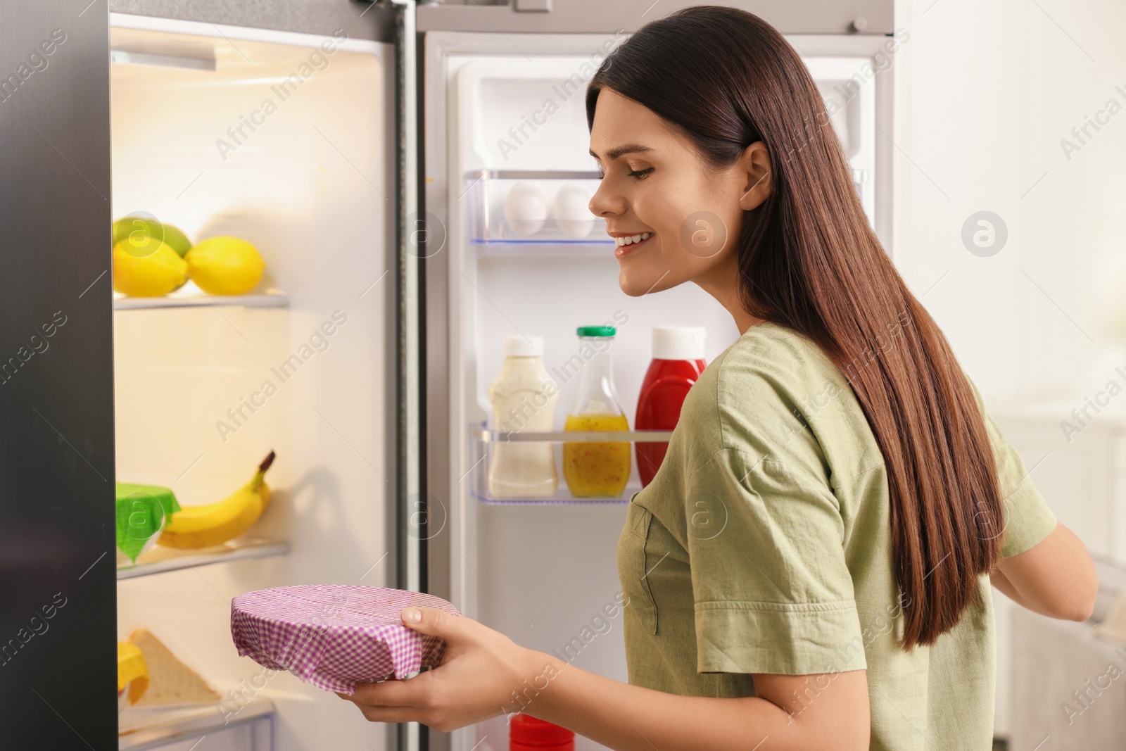 Photo of Happy woman holding bowl covered with beeswax food wrap near refrigerator in kitchen