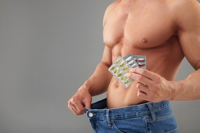 Photo of Weight loss. Athletic man with pills wearing big jeans on grey background, closeup. Space for text