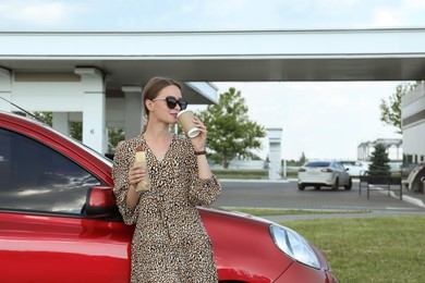 Photo of Beautiful young woman with hot dog drinking coffee near car at gas station