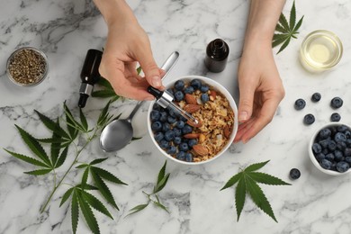 Photo of Top view of woman dripping THC tincture or CBD oil into oatmeal bowl at white marble table, closeup