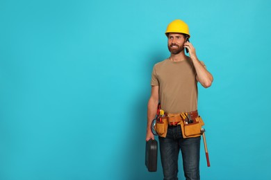 Photo of Professional builder in hard hat with tool belt talking on phone against light blue background, space for text