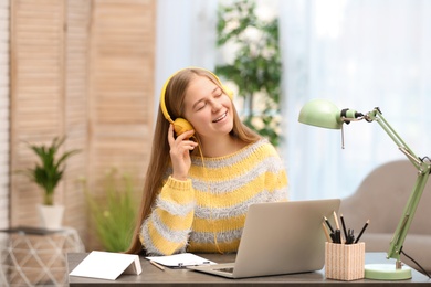 Pretty teenage girl listening to music with headphones at table in room