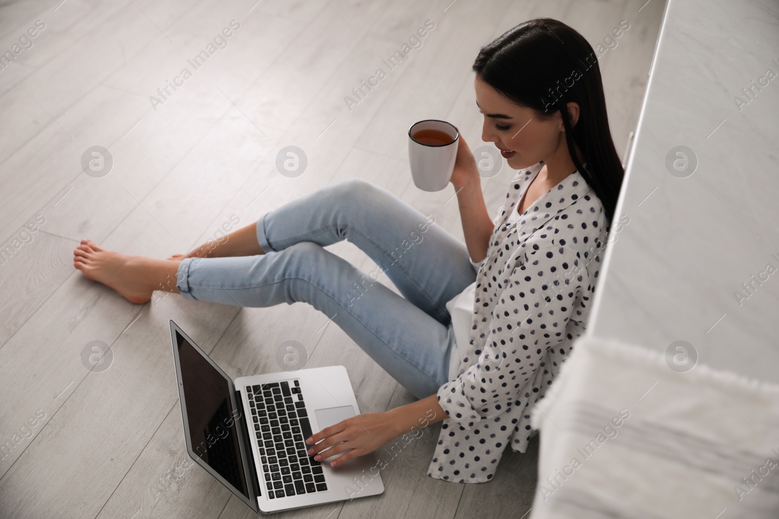 Photo of Happy woman with laptop and cup of drink sitting on warm floor indoors. Heating system