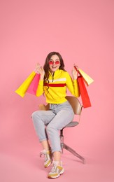 Photo of Emotional woman in stylish sunglasses holding many colorful shopping bags on armchair against pink background