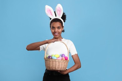 Photo of Happy African American woman in bunny ears headband holding wicker basket with Easter eggs on light blue background