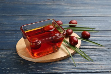 Photo of Palm oil in glass bowl with fruits and tropical leaf on blue wooden table