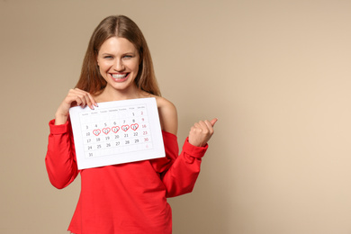 Photo of Excited young woman holding calendar with marked menstrual cycle days on beige background. Space for text