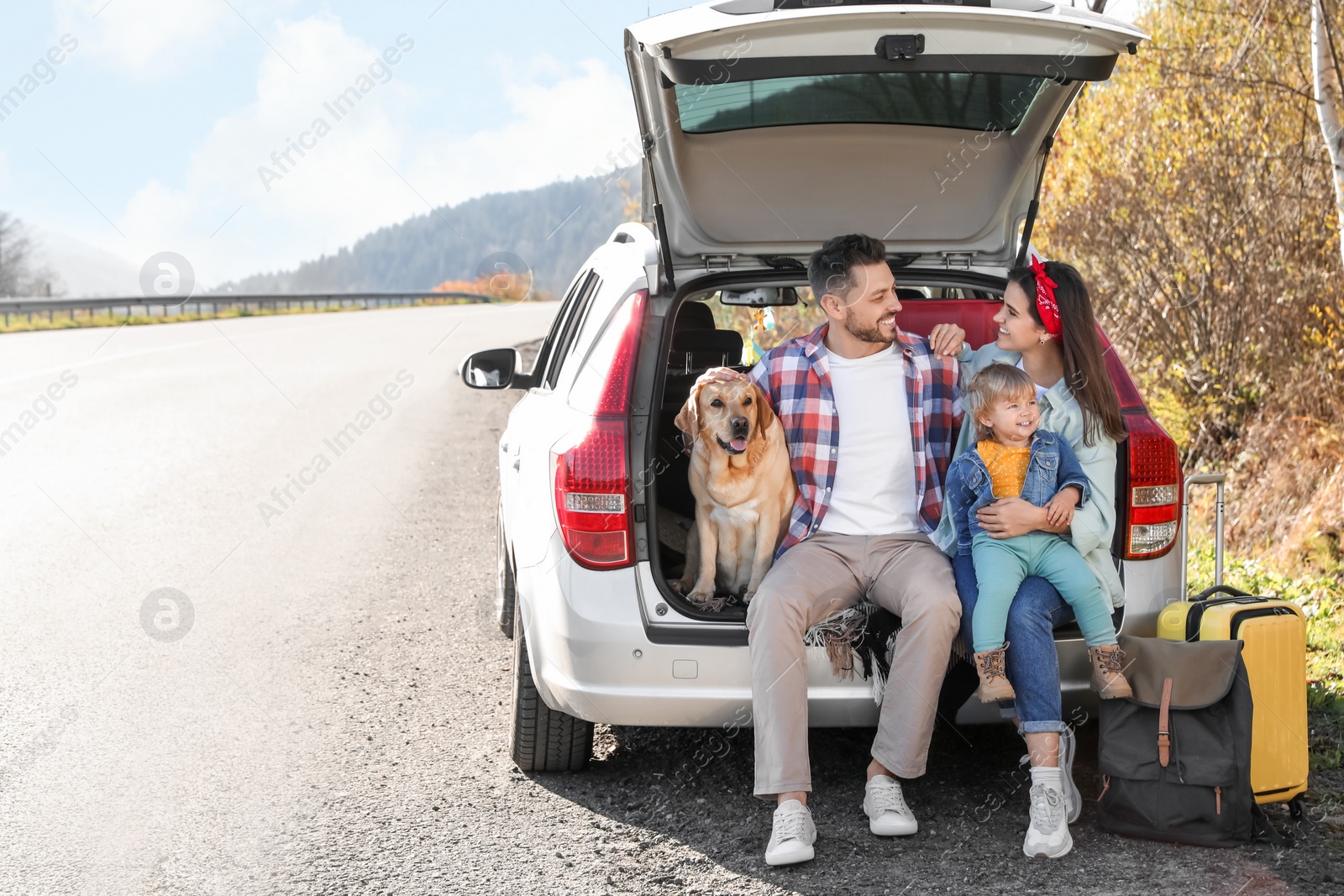 Photo of Parents, their daughter and dog sitting in car trunk near road, space for text. Family traveling with pet