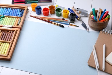 Photo of Blank sheet of paper, colorful chalk pastels and other drawing tools on white wooden table. Modern artist's workplace